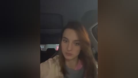 Media: A video of a young woman with long brown hair, fair skin, and a neutral expression, seated in a dimly lit car interior. She wears a beige jacket over a pink top. The background shows the car's dashboard and seats.