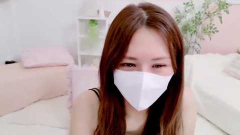 Media: Video of a young Asian woman with long brown hair, wearing a white medical mask, lying on a white bed in a pastel-colored room with soft lighting.