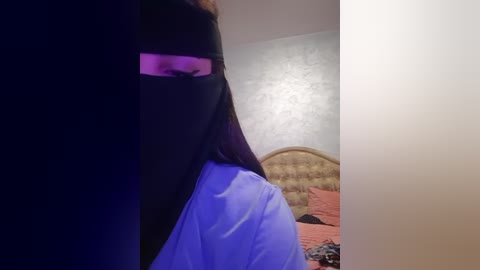 Media: A video of a person wearing a black niqab, revealing only the eyes, in a dimly lit bedroom with a patterned headboard and pink bedding.