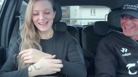 Media: Video of a smiling young woman with long blonde hair and a tattooed hand, sitting next to an older man with a tattooed arm, both wearing black hoodies in a car.