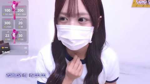 Media: A video of an East Asian woman with long brown hair, wearing a white short-sleeved shirt and a surgical mask, taken indoors with a simple white background.