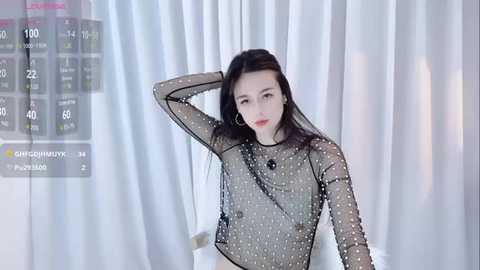 Media: Video of an Asian woman with long black hair, fair skin, wearing a sheer black top with polka dots, standing against white curtains.