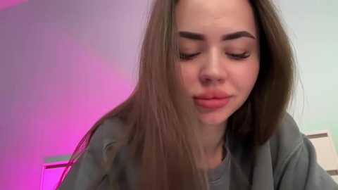 Media: Video of a young woman with long, straight brown hair, closed eyes, and a neutral expression, wearing a gray shirt, set against a softly lit, blurred background with pink and purple hues.