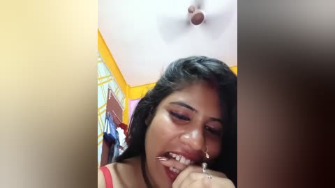 Media: Video of a young South Asian woman with long black hair and dark skin, wearing a red top, smiling and biting her finger, standing in a brightly lit room with yellow walls and a ceiling fan.