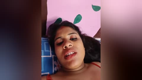 Media: Video of a young, dark-skinned woman with curly hair lying on a blue bedspread, wearing a red top, with a pink wall and green leaf cutout in the background.