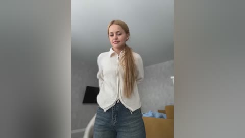 Media: Video of a young Caucasian woman with long blonde hair, wearing a white blouse and blue jeans, standing in a modern, minimalist room with white walls, a TV, and a wooden dresser.