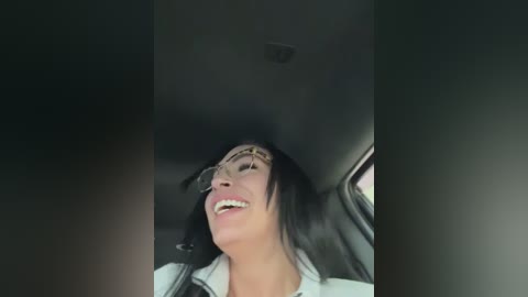 Media: Video of a smiling woman with dark hair and glasses, wearing a white shirt and black jacket, seated in a dimly lit car.