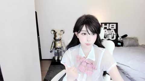 Media: A video of an East Asian woman with long black hair, fair skin, and a pale pink bowtie, sitting in a minimalist, white room with plush toys and a black desk chair.