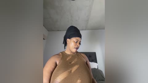 Media: Video of a plus-size Black woman with a medium brown complexion wearing a sheer brown top, black headscarf, and minimal makeup. She stands in a modern, minimalist bedroom with a grey headboard and white walls.