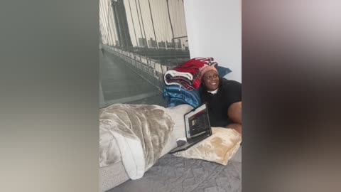 Media: Video of a happy Black woman with a medium build, wearing a black shirt, lying on a bed with a laptop, surrounded by neatly folded blankets. The background features a large wall mural of a bridge.