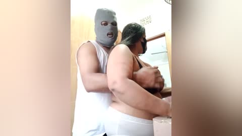 Media: Video of a muscular man wearing a balaclava, holding a large-breasted woman in a white tank top, in a dimly lit bathroom with beige walls and a showerhead.