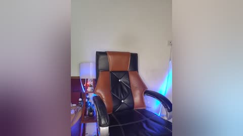 Media: A video of a brown leather and black vinyl gaming chair with a tufted backrest, situated in a cluttered room with a white wall and a wooden desk with a lamp and scattered items.