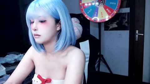 Media: A video of a young woman with fair skin and shoulder-length, pastel blue hair, wearing a strapless red and white dress, sitting in a dimly lit room. A colorful pinball machine and posters adorn the background.