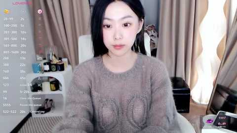 Media: A video of an Asian woman with long black hair, wearing a gray knit sweater, sitting in a cluttered room with beige curtains, a TV, and various objects.