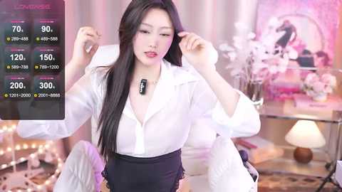 Media: Video of an Asian woman with long black hair, wearing a white blouse and black skirt, adjusting her hair. Background includes a pink floral arrangement and a lamp.