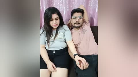 Media: Video of a South Asian couple, a woman in a grey top and black skirt, and a man in a pink t-shirt and black pants, sitting close, with a pink curtain in the background.