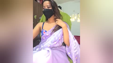 Media: Video of a young woman with medium brown skin, wearing a purple sari with floral patterns, black mask, and blue bra, sitting on a green chair in a dimly lit room with fairy lights.