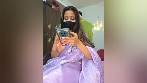 Media: Video of a woman in a lavender sari with floral embroidery, wearing a black face mask, taking a selfie with a blue phone, in a modern, softly lit room.