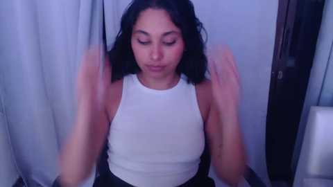 Media: Video of a young woman with medium skin tone and shoulder-length dark hair, wearing a white sleeveless top, sitting on a bed with white curtains in the background.