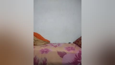 Media: Video of a simple bedroom with a bed covered in a floral-patterned pink and purple bedspread, featuring a beige pillow on the left and an orange pillow on the right. The walls are plain white.