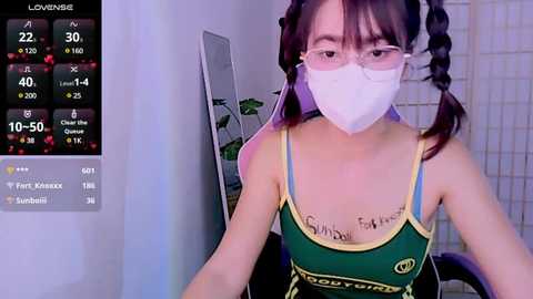 Media: Video of an East Asian woman with long black hair in twin braids, wearing a green tank top with yellow accents, a white surgical mask, and glasses, sitting in a dimly lit room with a monitor showing live stream stats.