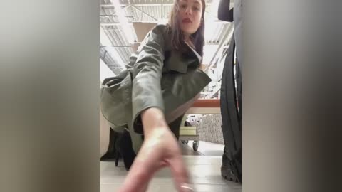 Media: A video of a woman in a grey coat, crouching and reaching forward, in a dimly lit, cluttered room with wooden floors and exposed ceiling beams.