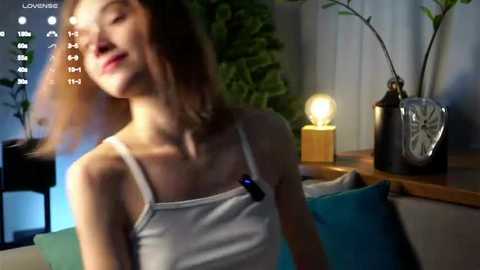 Media: Video of a young woman with shoulder-length brown hair, wearing a white tank top, standing in a dimly lit room with plants, a lit candle, and a clock.