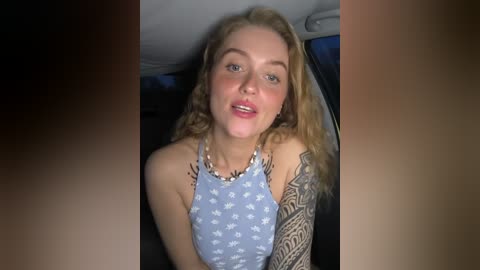 Media: Video of a young, fair-skinned woman with curly blonde hair, wearing a blue sleeveless top, seated in a car. She has a large tattoo on her left arm.