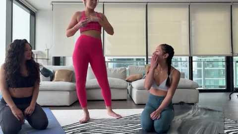 Media: Video of three women in a modern living room. Two women in sportswear, one in a pink bra and high-waisted leggings, pose confidently. The third woman, kneeling, covers her face with her hands, laughing.