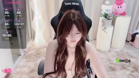 Media: Video of a young Asian woman with long brown hair, wearing a black gaming chair with the name \"Tessa Bland\" on it, sitting in a cozy, pink carpeted room with a plush pink pillow and a white table with a plush toy.