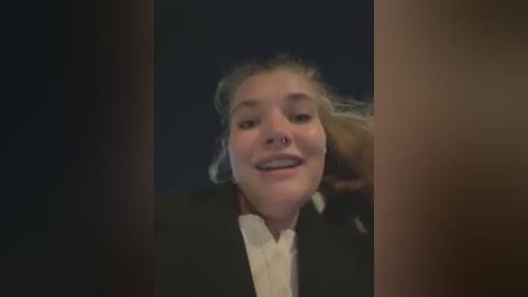 Media: Video of a young woman with fair skin and light brown hair, smiling with a nose ring. She wears a black blazer over a white shirt. The background is dark and blurry.