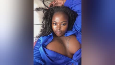 Media: Video of a Black woman with dark skin, wearing a blue shirt unbuttoned to reveal her large breasts, lying on a bed with white sheets, hands in her hair.