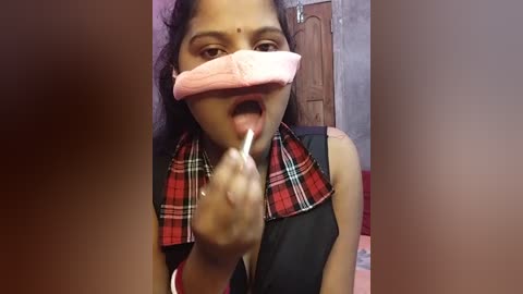 Media: Video of an Indian woman with medium skin tone, wearing a plaid scarf, gagged with pink tape, and giving a middle finger in a room with a wooden door and red walls.