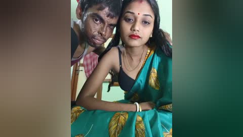 Media: Video of a young woman in a teal sari with leaf patterns, wearing a red bindi, sitting next to a man with a white substance on his face, both smiling.