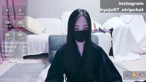 Media: A video of a young East Asian woman with long black hair, wearing a black face mask, sitting on a couch in a dimly lit living room.