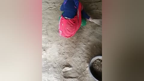 Media: Video of a toddler wearing a blue shirt and red skirt playing in a sandbox, surrounded by light-colored sand, with a small plant pot in the corner.