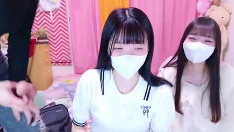 Media: Video of two young Asian women with long black hair, wearing white masks, in a pink room with a yellow door and patterned wallpaper, holding bags.