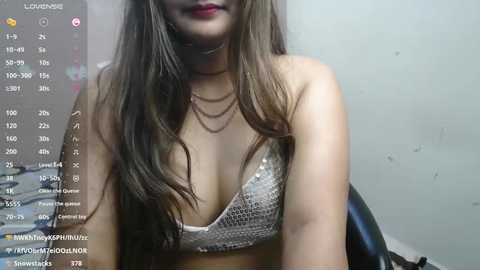 Media: A video of a young woman with long brown hair, wearing a silver sequined bra, and a layered necklace. The background is a plain white wall with a live stream overlay displaying time, date, and social media handles.