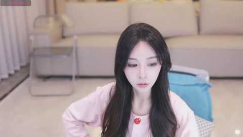 Media: Video of an East Asian woman with long black hair and fair skin, wearing a pink sweater, sitting on a light-colored rug in a modern living room with beige couches, a blue laundry basket, and a metallic side table.