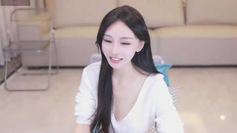 Media: Video of a smiling, fair-skinned Asian woman with long black hair, wearing a white top, sitting on a cream-colored sofa in a modern living room.