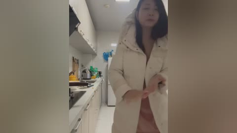 Media: A candid video of an Asian woman with long black hair, wearing a white coat, adjusting her clothing in a modern kitchen with white cabinets and stainless steel appliances.