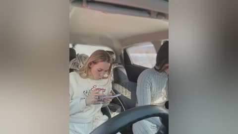 Video of a blonde woman in a white sweater reading a newspaper, sitting in a car with a grey-haired man in a striped sweater in the background.