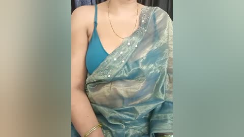 Media: Video of a South Asian woman wearing a turquoise saree with a silver blouse, a gold necklace, and bangles. The background is a plain beige wall with a dark curtain.