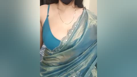 Media: Video of a woman with medium skin tone, wearing a blue blouse and a shimmering green sari with intricate embroidery, her black hair visible, indoors, wearing a mask.