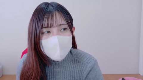 Media: Video of an Asian woman with long, straight brown hair, wearing a grey turtleneck sweater, white face mask, and red shirt, sitting indoors with a plain white wall background.