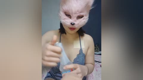 Media: Video of a young Asian woman in a furry, pink fox mask, with medium-length black hair, wearing a grey lace bra, holding a water bottle, in a dimly lit room.