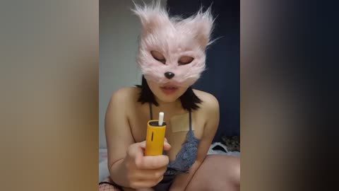 Media: Video of a young Asian woman in a blue lace bra, wearing a fluffy, pink fox mask, lighting a yellow lighter. She has shoulder-length black hair. Background is dark, blurry.