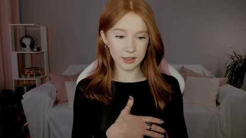 Media: Video of a pale-skinned, red-haired young woman with long hair, wearing a black top, speaking on a smartphone in a cozy, dimly lit living room with white furniture, shelves, and a plant.