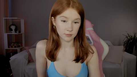 Media: Video of a young, fair-skinned, red-haired woman with a slender physique, wearing a blue tank top, sitting in a dimly-lit, cozy living room with pastel furniture and a pink blanket.