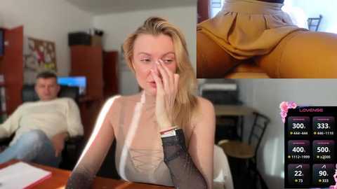 Media: A video captures a blonde woman with a fair complexion and medium build, wearing a sheer black top and a yellow skirt. She covers her mouth with her hand, appearing thoughtful. The background shows a room with a man in a white sweater and a TV.